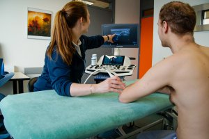 Een fysiotherapeute wijst op een scherm aan wat door echografie onderzocht wordt terwijl de echokop bij een mannelijke patiënt op zijn pols is geplaatst. 