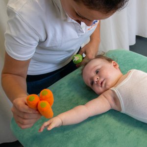Een foto uit een manuele fysiothearpie sessie waar een fysiotherapeute een knuffel naast een klein kindje houdt waardoor het kind zijn arm uitreikt. 