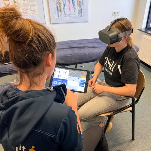 Een fysiotherapeute zit op een Ipad terwijl haar patiënt een Virtual Reality training ondergaat. 