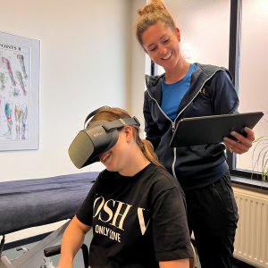 Een fysiotherapeute kijkt lachend naar haar Ipad terwijl haar patiënt lachend met een VR bril de virtual reality training ondergaat