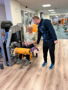 Een vrouwelijke 55+'er zit op een sport apparaat in een fitness ruimte waarbij ze wordt ondersteund door een medewerker van Medifit